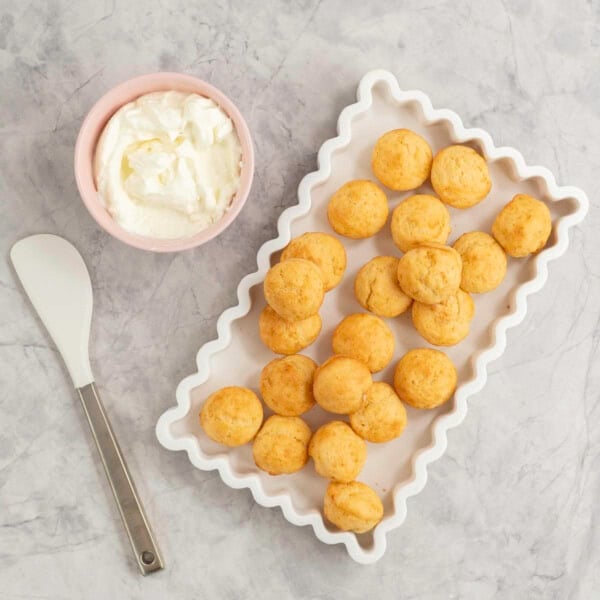 White rectangle dish with wavy border, batch of mini muffins inside, small pink ramekin with greek yogurt inside and a spatula on bench top.