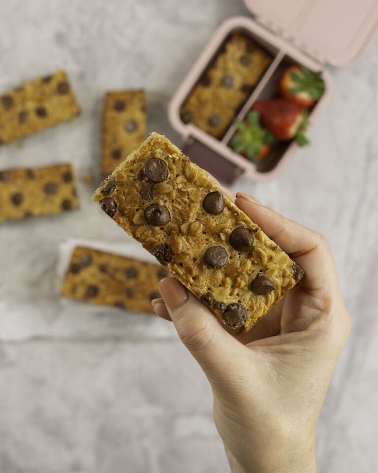 Woman's hand holding a golden granola bar with lunchbox below.