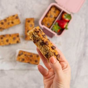 Woman's hand holding a golden granola bar with lunchbox below.