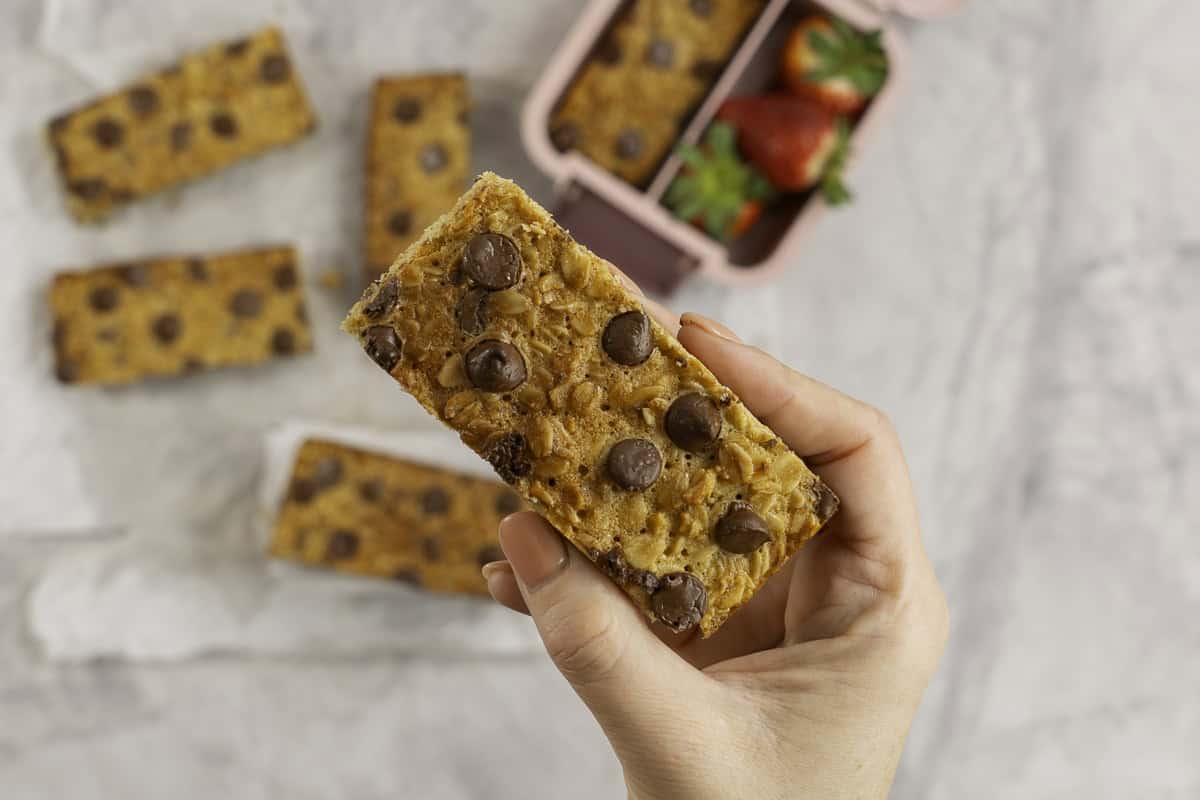 Woman's hand holding a golden granola bar with lunchbox below.