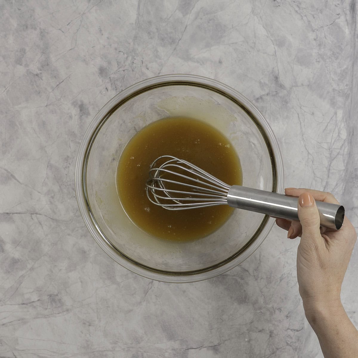 Hand holding a whisk with sugar and butter mixture in a glass bowl on benchtop.