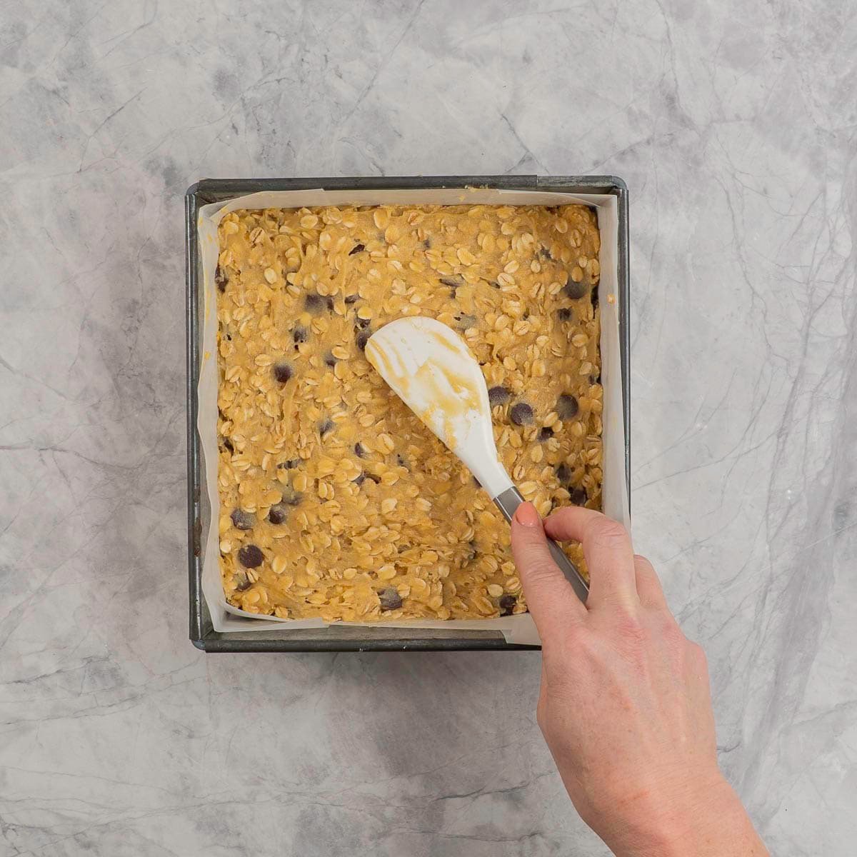 Granola bar mixture being pressed with spatula into a square lined tin.