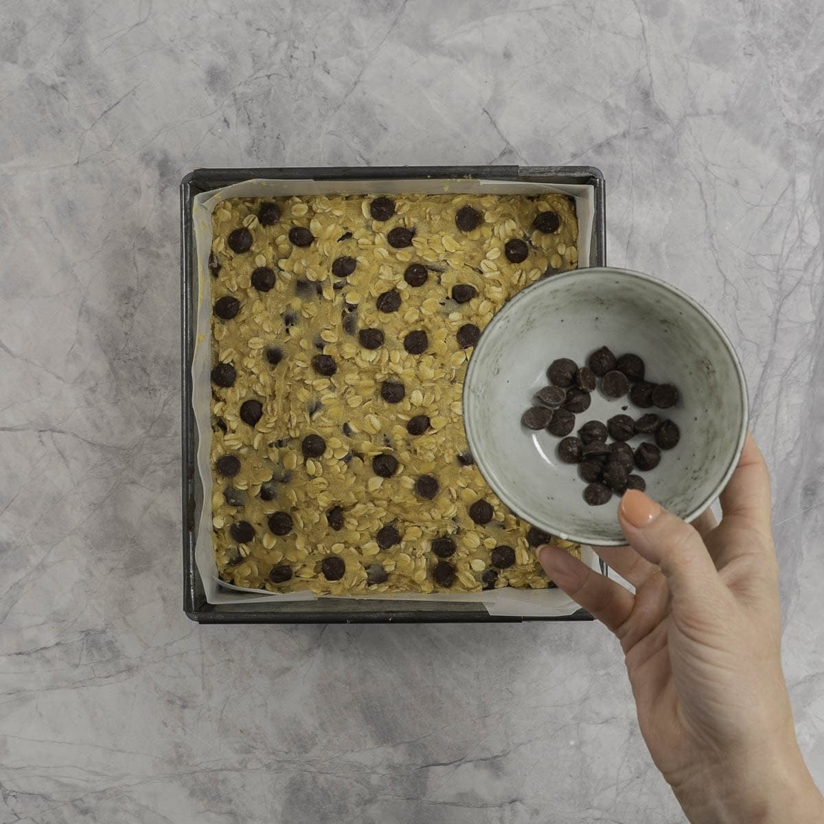 Granola bar mixture in a square lined tin with chocolate chips on top and woman's hand holding a bowl with chocolate chips inside.