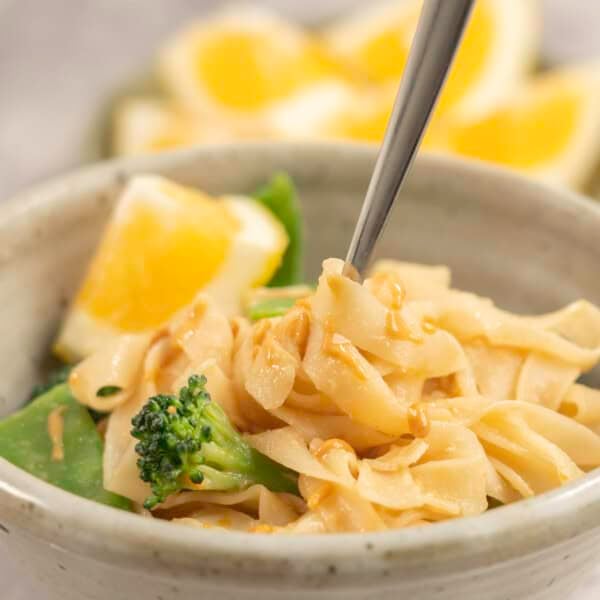 Dinner bowl on benchtop with rice noodles with peanut butter mixture added throughout, broccoli and snow heads inside and garnished with slice of lemon.