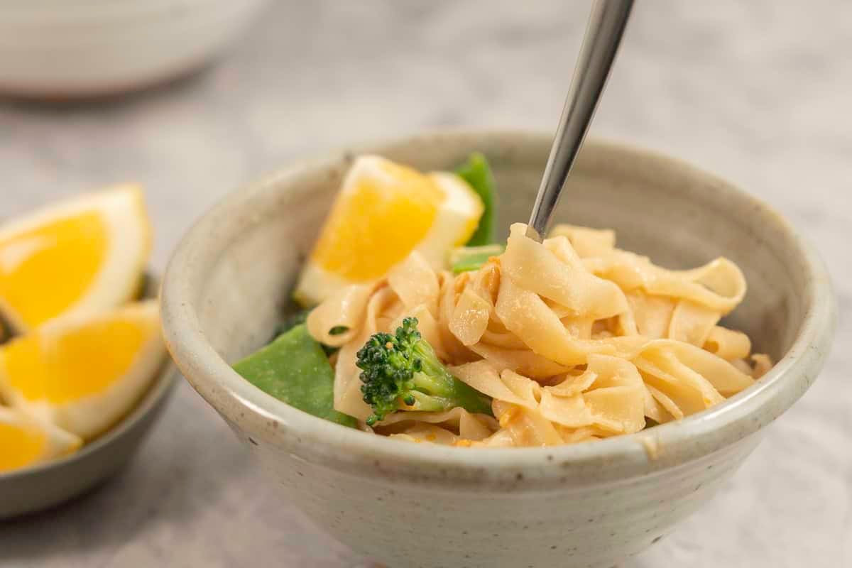 Dinner bowl on benchtop with rice noodles with peanut butter mixture added throughout, broccoli and snow heads inside and garnished with slice of lemon.