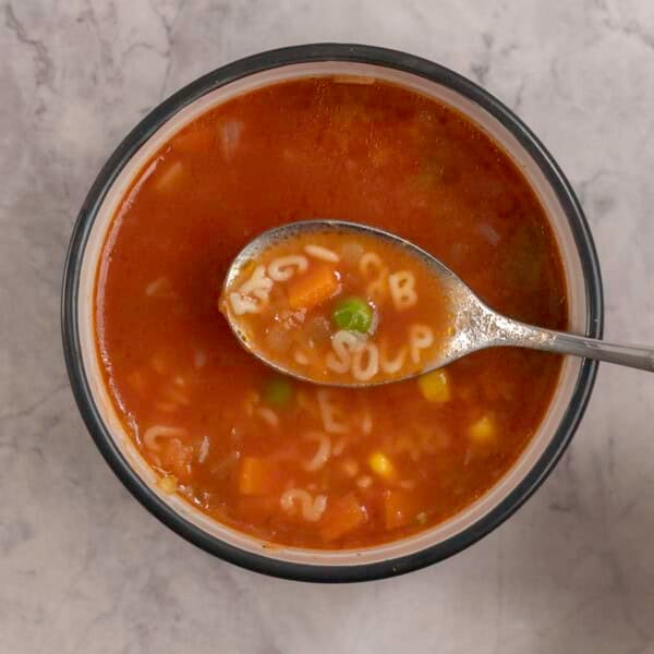 Woman's hand holding a scoop of soup on spoon with pasta letters spelling "soup", bowl with soup inside on benchtop below.