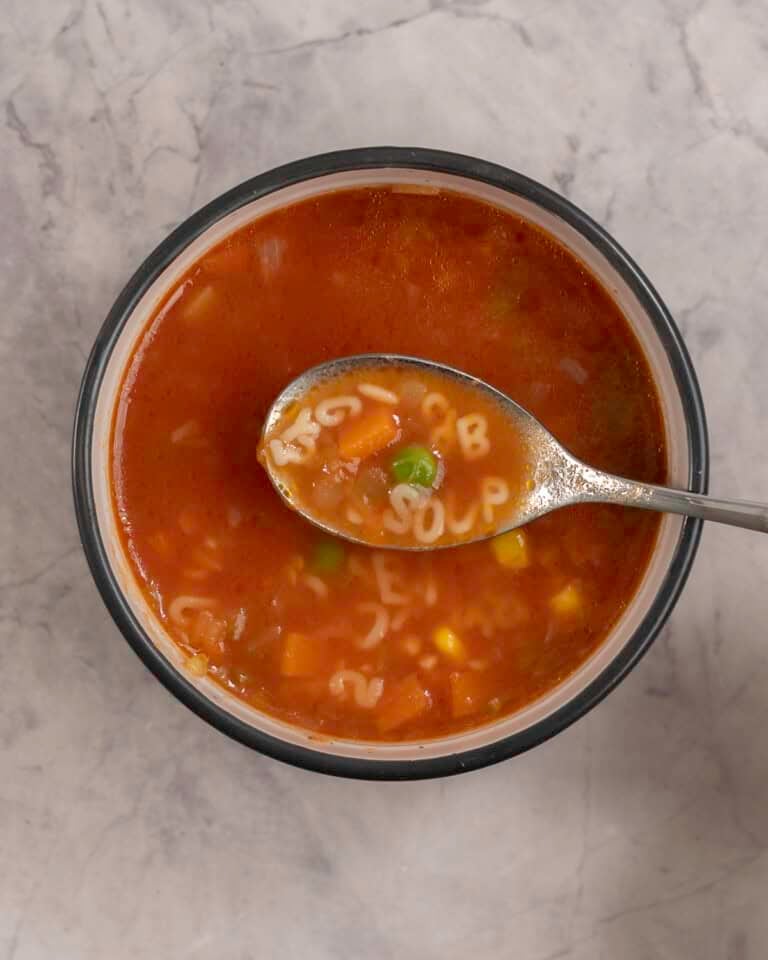 Woman's hand holding a scoop of soup on spoon with pasta letters spelling "soup", bowl with soup inside on benchtop below.