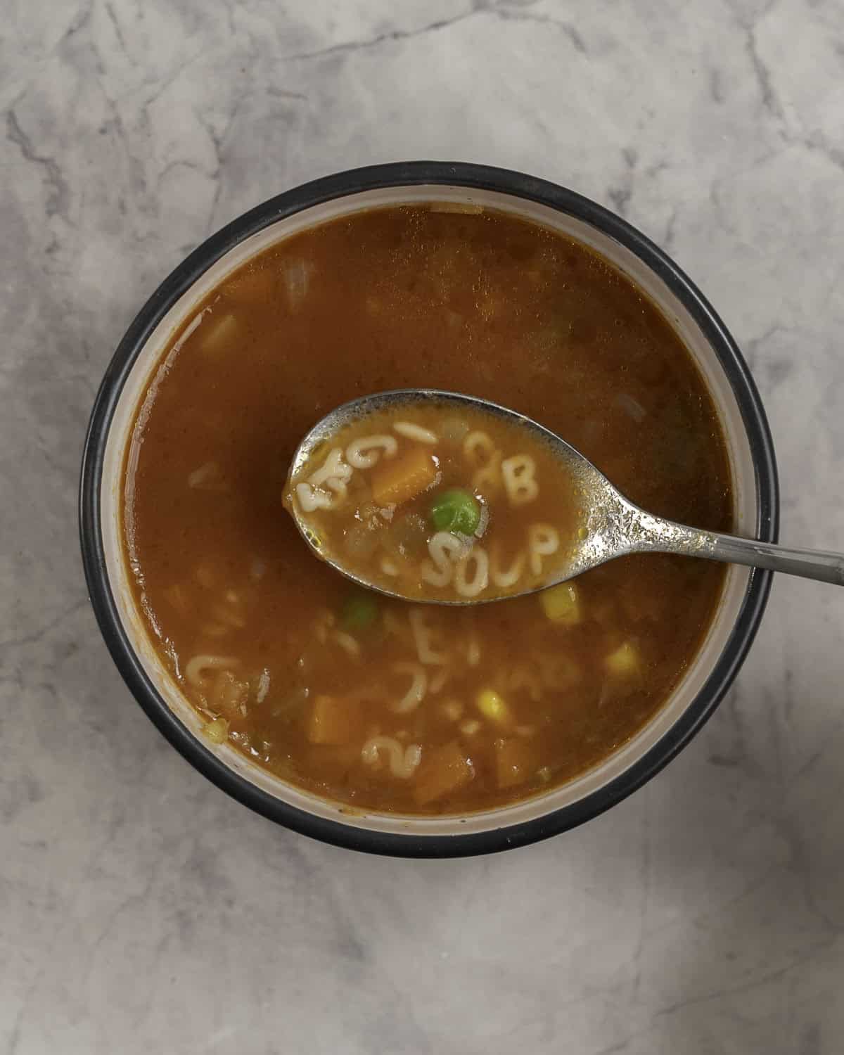 Woman's hand holding a scoop of soup on spoon with pasta letters spelling "soup", bowl with soup inside on benchtop below.