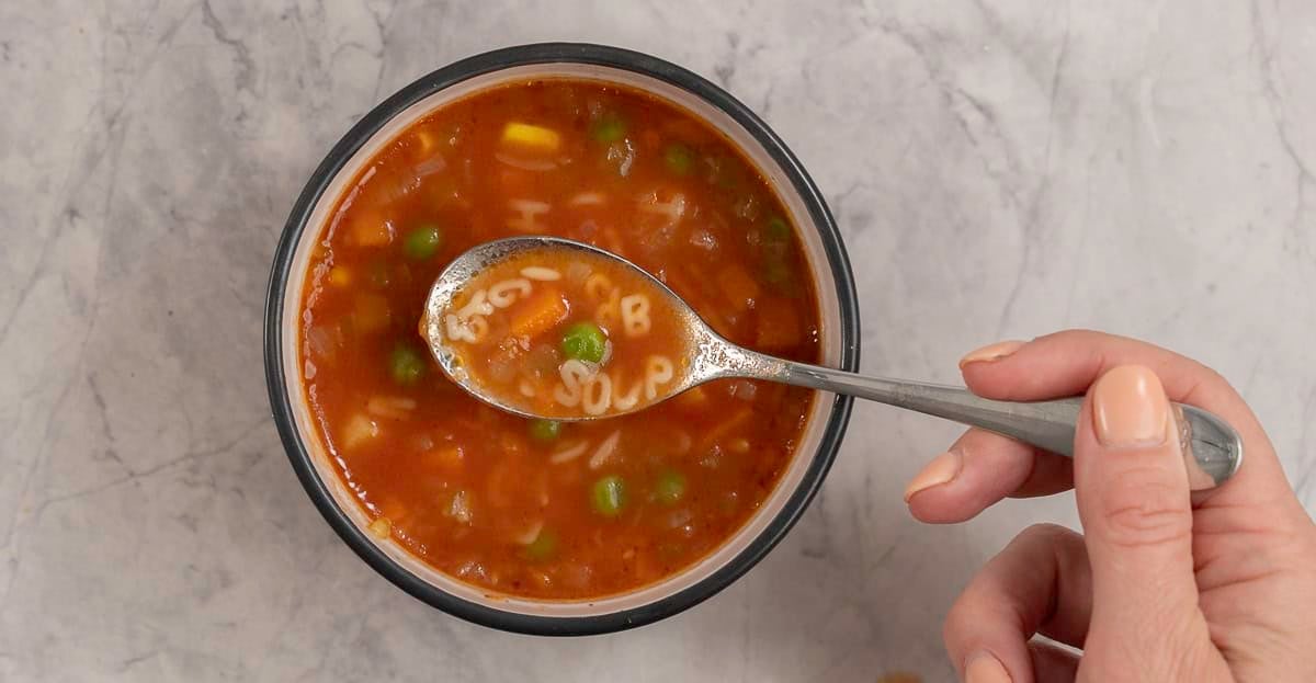 Woman's hand holding a scoop of soup on spoon with pasta letters spelling "soup", bowl with soup inside on benchtop below.