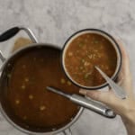 Woman's hand holding a small bowl with spoon inside and vegetable soup with alphabet pasta, pot of soup below on benchtop with a ladle.