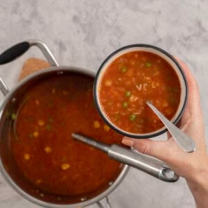 Woman's hand holding a small bowl with spoon inside and vegetable soup with alphabet pasta, pot of soup below on benchtop with a ladle.