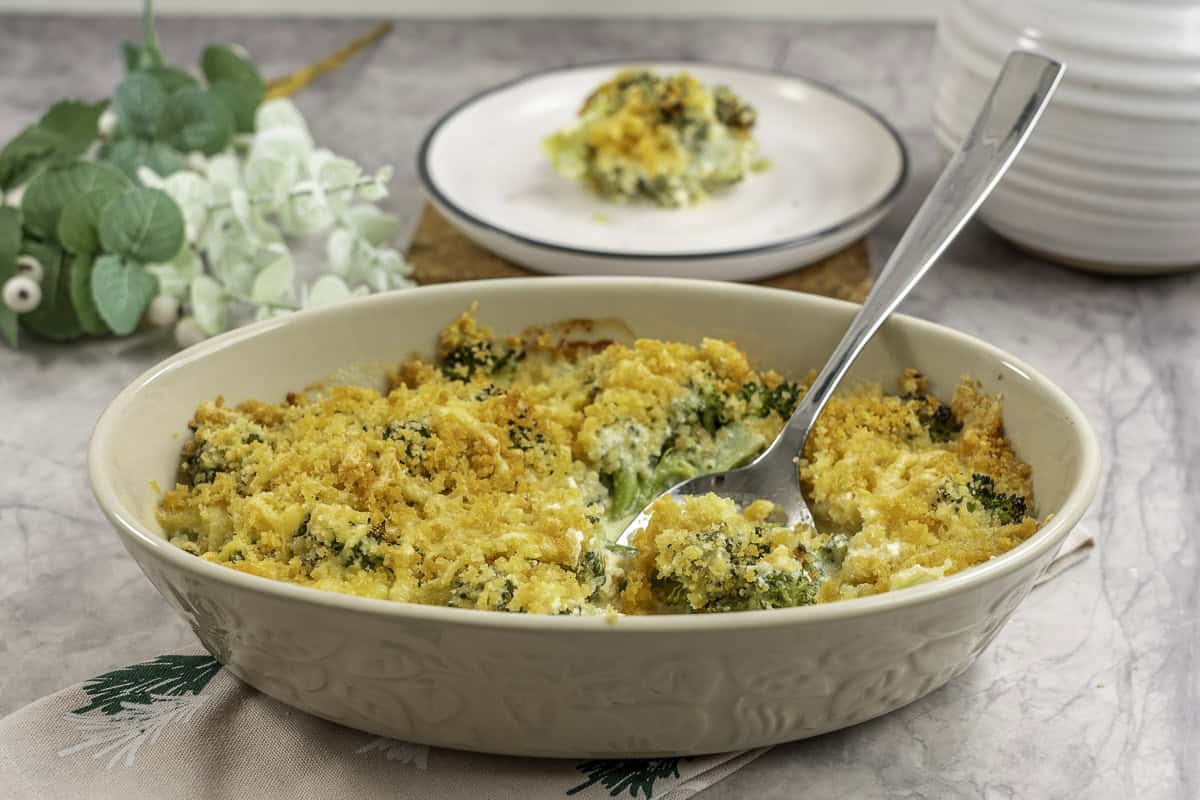 Broccoli cheese dish with crispy golden brown topping, metal spoon scoooping out a portion from inside oval casserole dish, plate with serving in background.