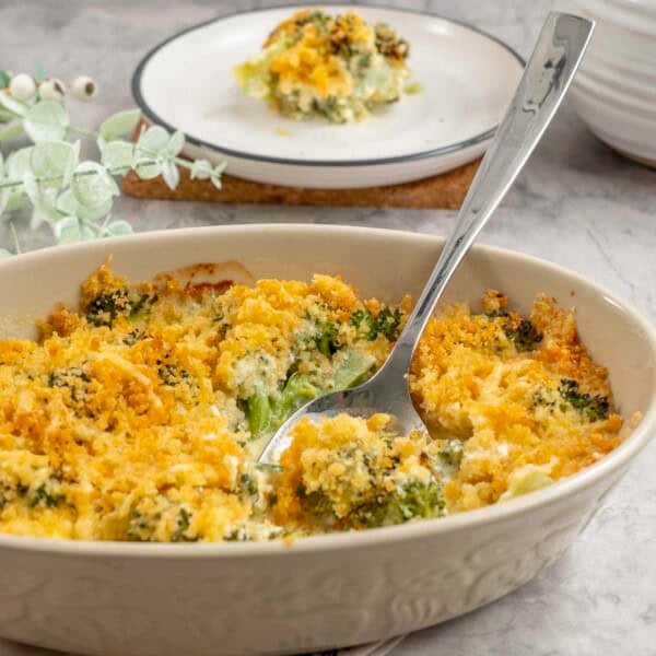 Broccoli cheese dish with crispy golden brown topping, metal spoon scoooping out a portion from inside oval casserole dish, plate with serving in background.