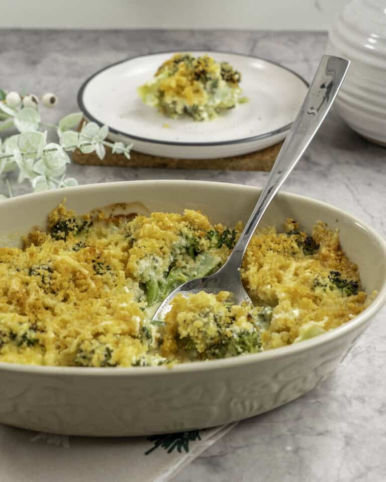 Broccoli cheese dish with crispy golden brown topping, metal spoon scoooping out a portion from inside oval casserole dish, plate with serving in background.