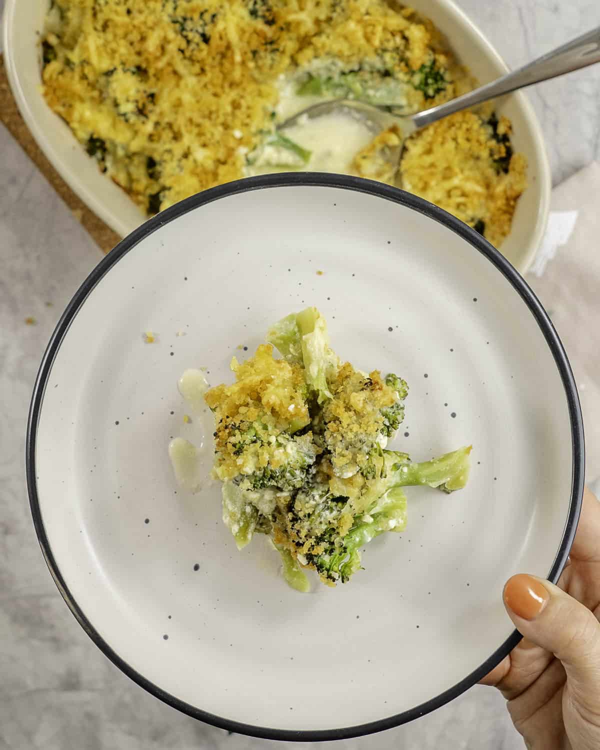 Womans hand holding a a dinner plate with portion of broccoli cheese, with golden brown toppings.