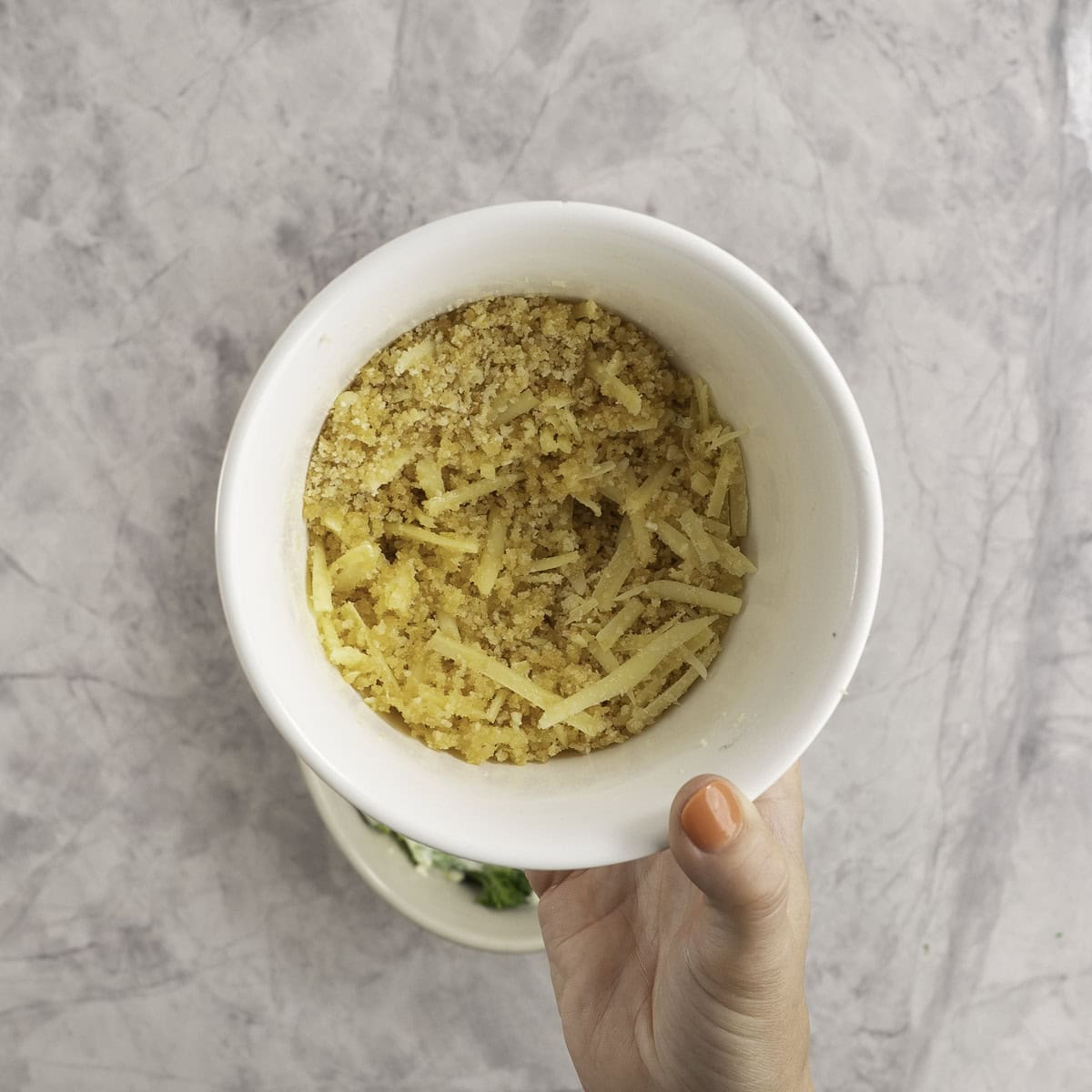 Womans hand holding a white bowl with breadcrumbs and grated cheese inside.