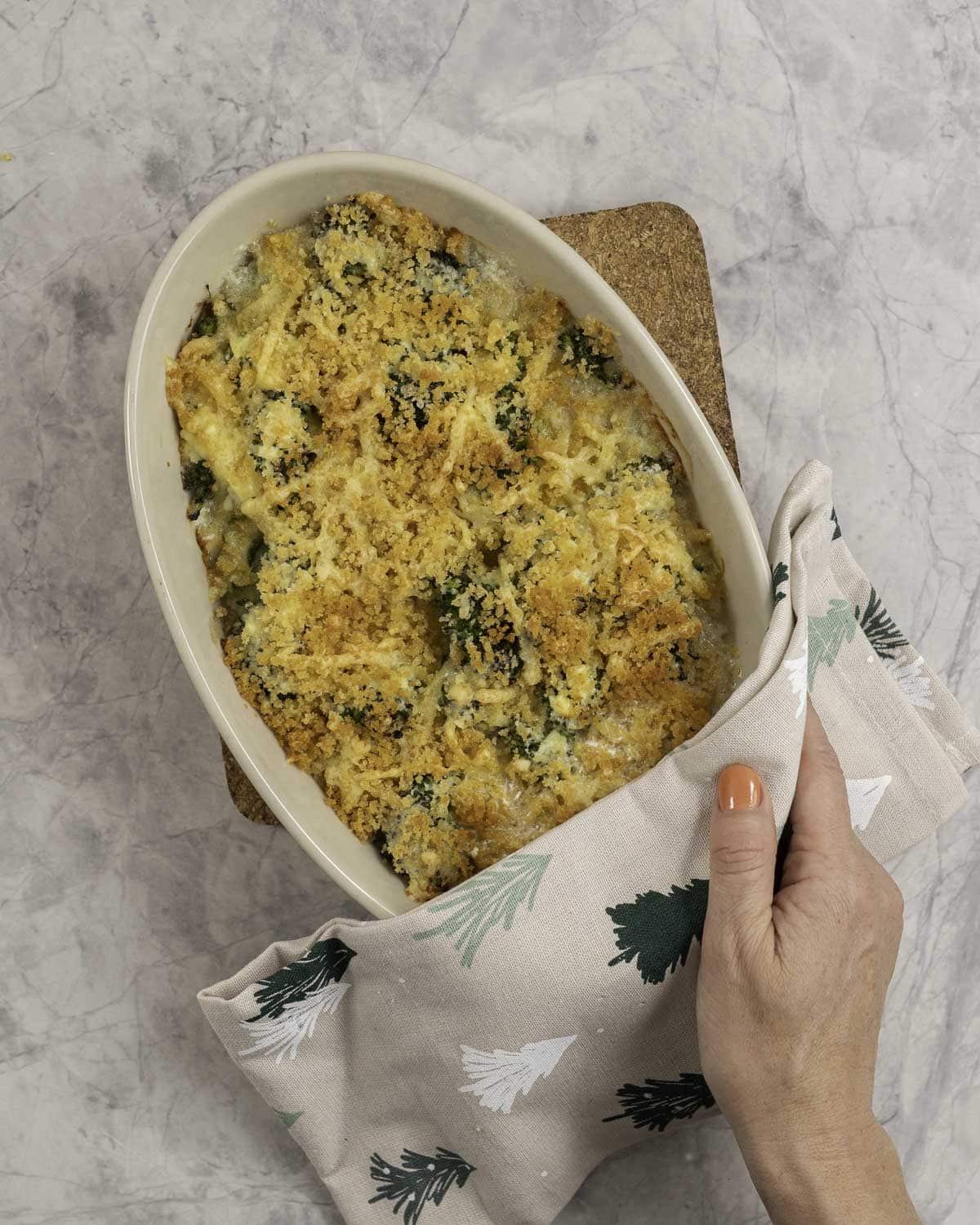Womans hand holding the edge of a casserole dish with a christmas tea towel, with golden brown breadcrumb toppings on broccoli inside.