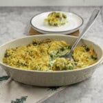 Broccoli cheese dish with crispy golden brown topping, metal spoon scoooping out a portion from inside oval casserole dish, plate with serving in background.