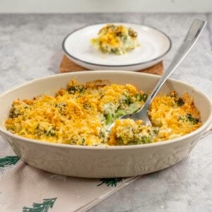 Broccoli cheese dish with crispy golden brown topping, metal spoon scoooping out a portion from inside oval casserole dish, plate with serving in background.