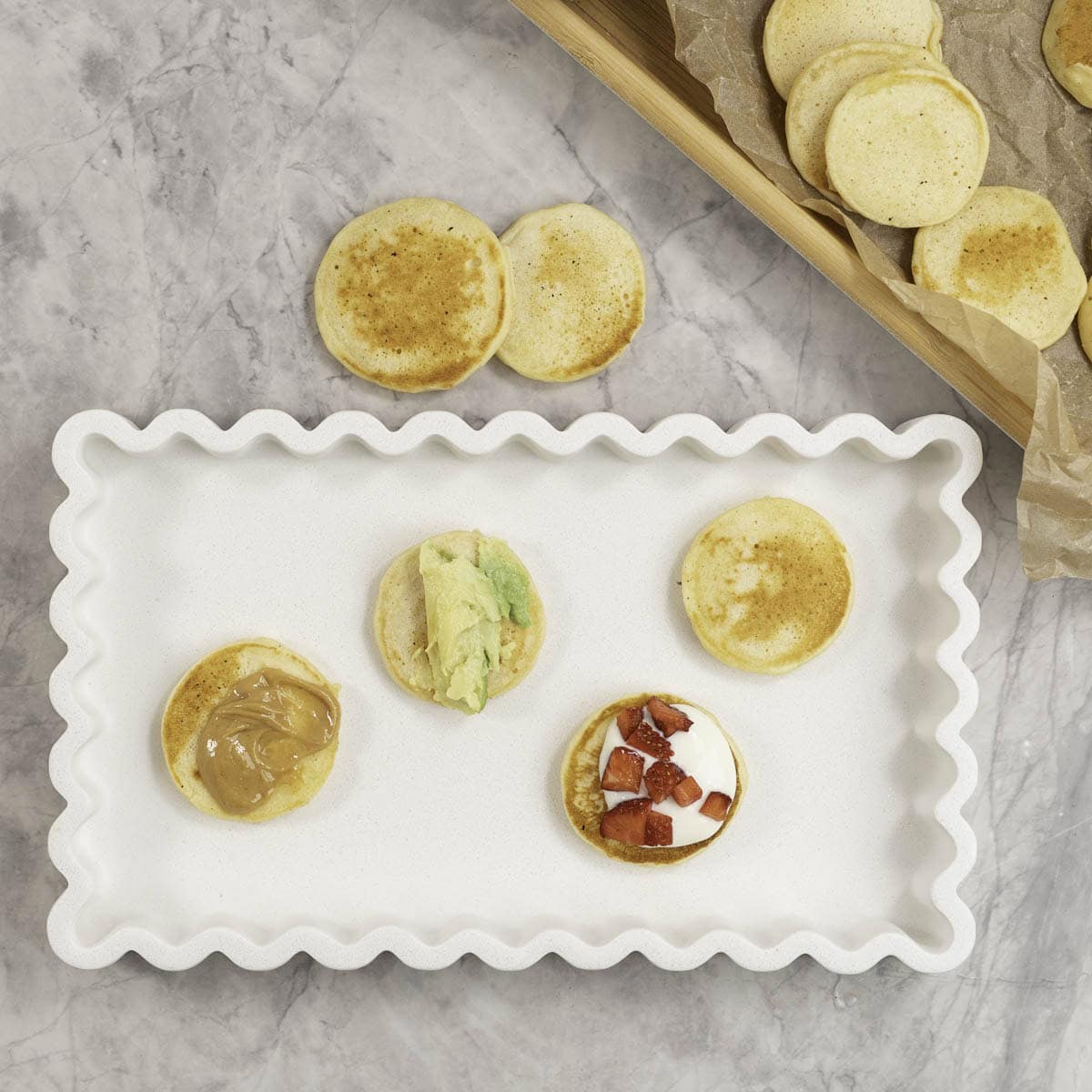 Four mini pancakes on a white rectangular serving dish, each differently topped with peanut butter, avocado, yogurt and strawberry, and one plain.