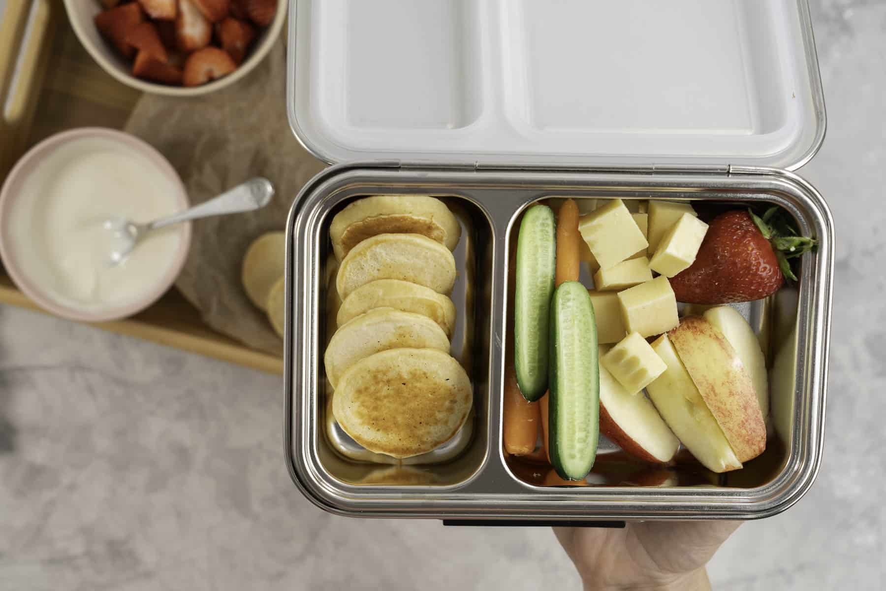 Hand holding a stainless steel lunchbox, one compartment with a row of mini pancakes, other with sliced apple, cucumber, carrot, diced cheese and a strawberry.