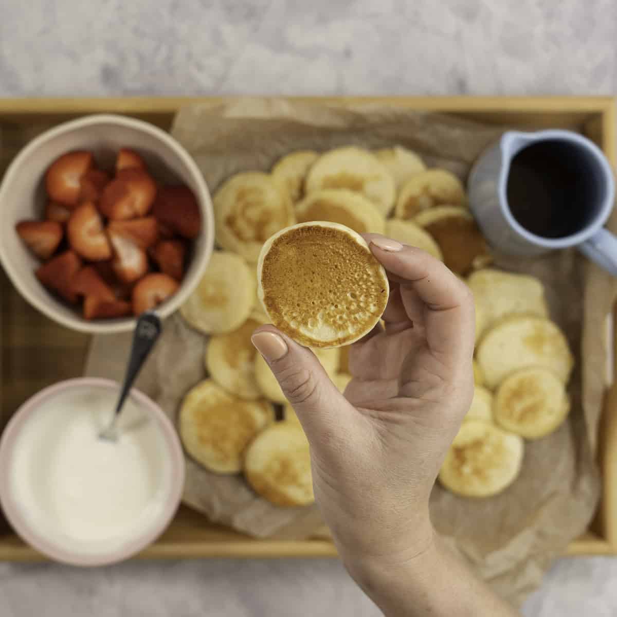 Womans hands holding a mini pancake, below on a tray lined with baking paper a spread of pancakes, a small jug of maple syrup, bowl of yogurt and cut strawberries.
