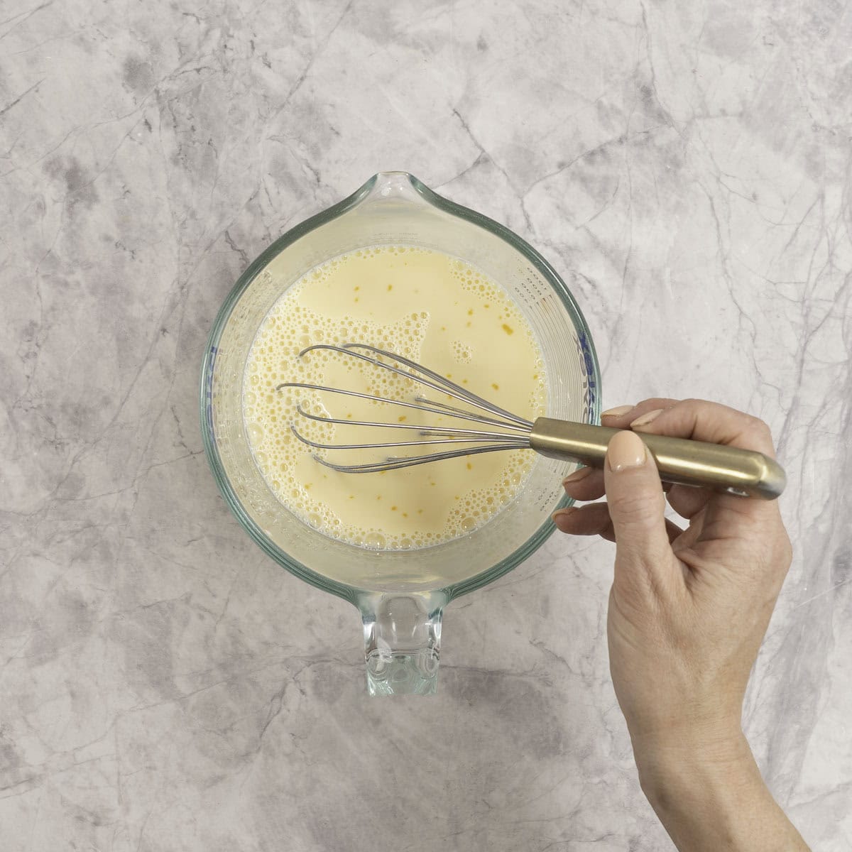 Wet ingredients inside a glass mixing jug with woman's hand mixing with a whisk.