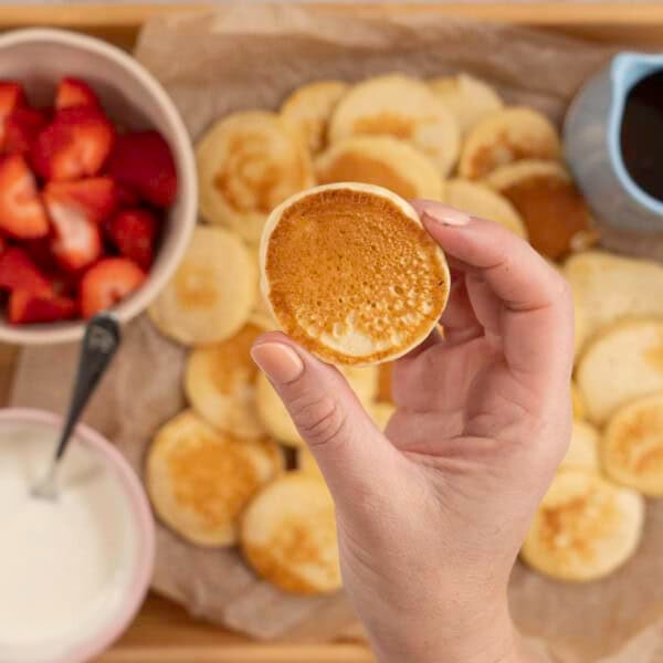 Womans hands holding a mini pancake, below on a tray lined with baking paper a spread of pancakes, a small jug of maple syrup, bowl of yogurt and cut strawberries.