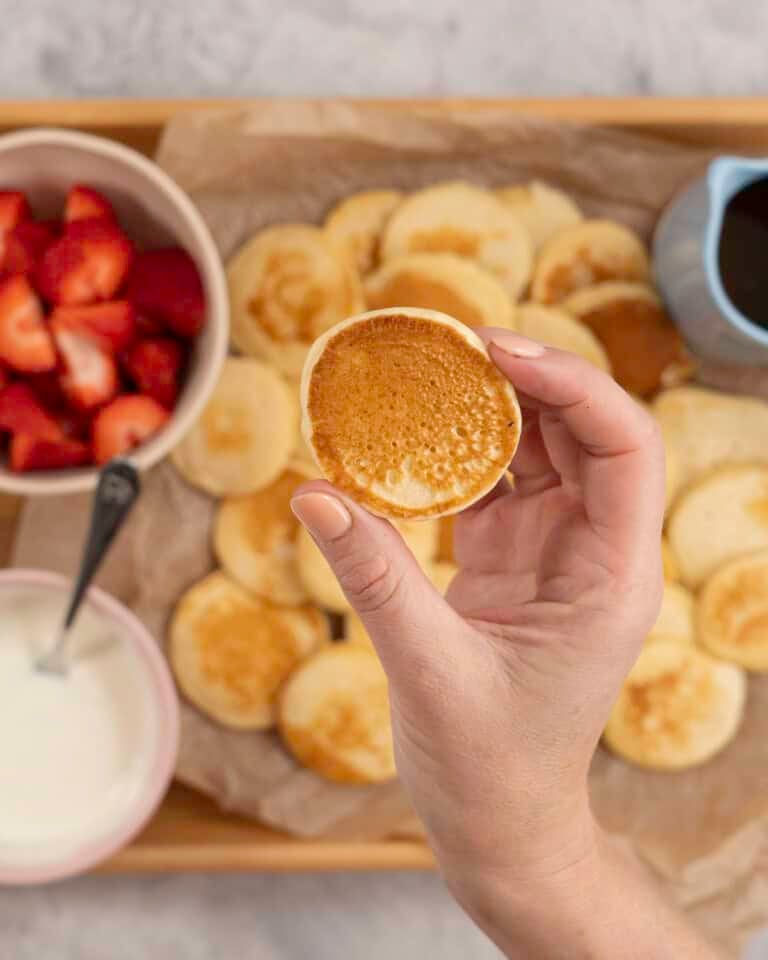 Womans hands holding a mini pancake, below on a tray lined with baking paper a spread of pancakes, a small jug of maple syrup, bowl of yogurt and cut strawberries.