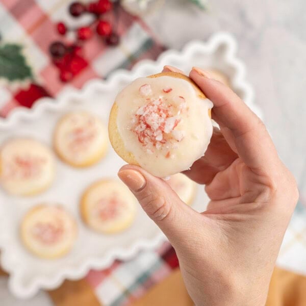 Woman's hand holding a small cookie with icing and crushed candy cane pieces on top, above a white decorative tray with more cookies inside below.