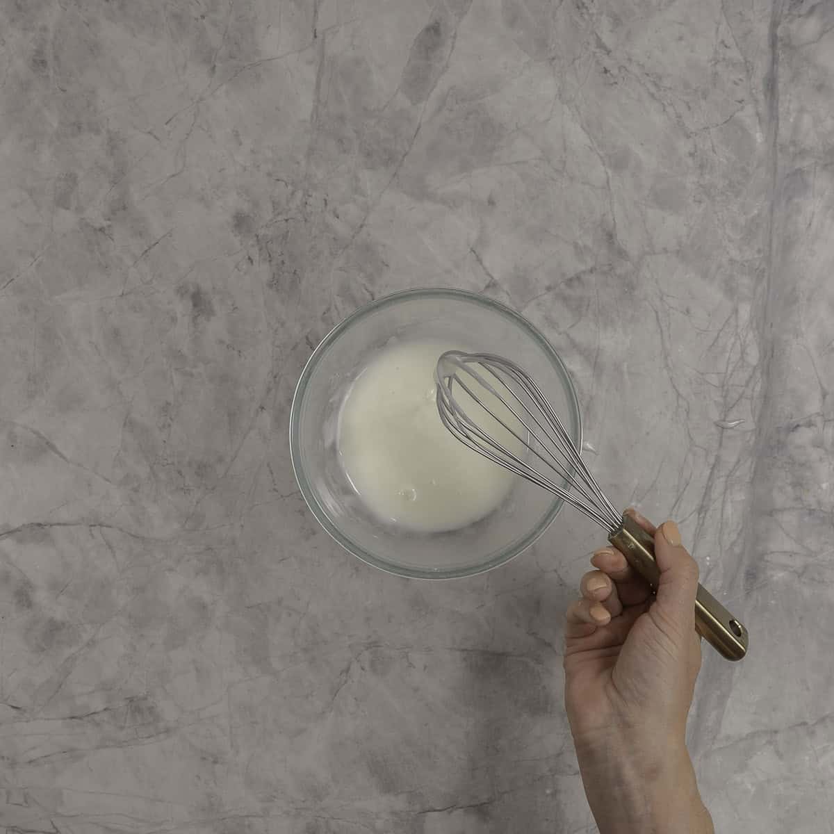 Womans hand holding a whisk stirring icing in a small glass bowl.