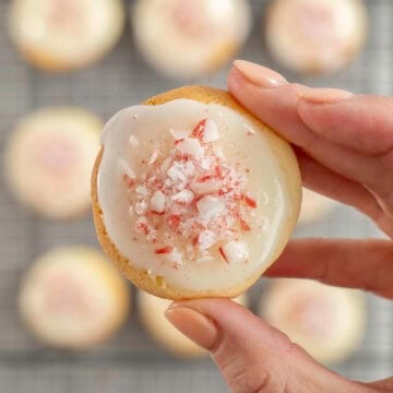 Woman's hand holding a small cookie with icing and crushed candy cane pieces on top, above a cooling tray with more cookies below.