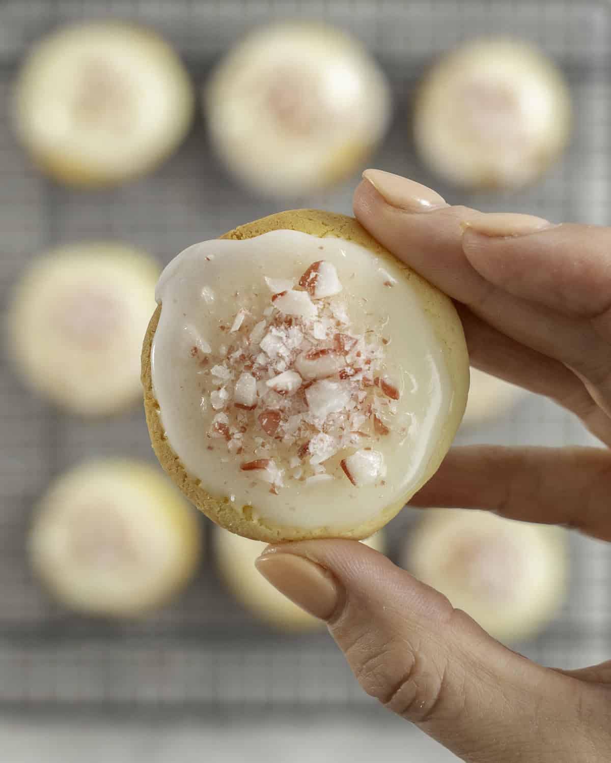 Woman's hand holding a small cookie with icing and crushed candy cane pieces on top, above a cooling tray with more cookies below.