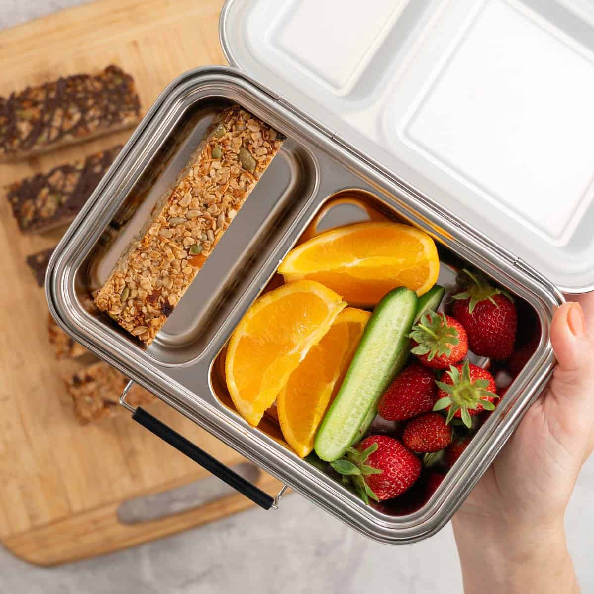 Woman holding a stainless steel lunchbox, one compartment with a homemade granola bar, and an assortment of fruit.