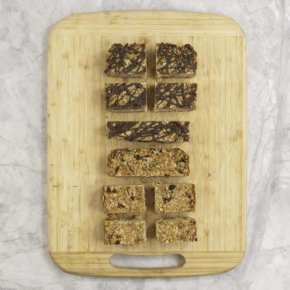 A wooden chopping board on marble bench top with cubes and sliced granola bars, three with melted chocolate drizzled on top.