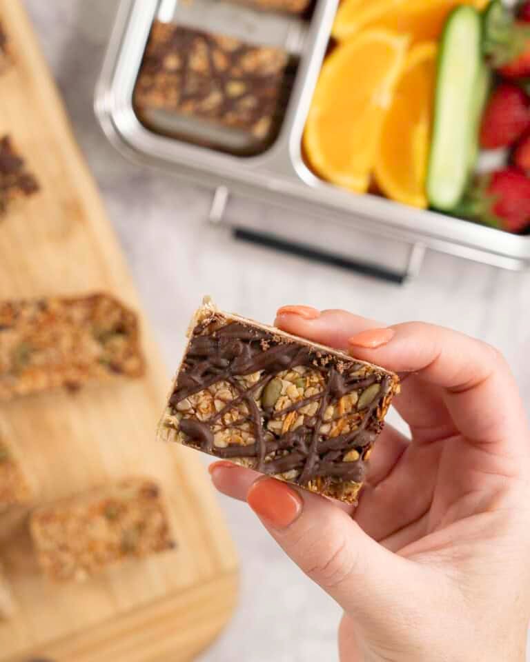 Womans hand holding a small square granola bar with drizzled melted chocolate.