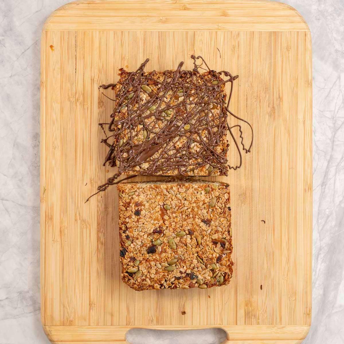 Wooden chopping board on benchtop with granola bar cut in half, one square drilled with melted chocolate.