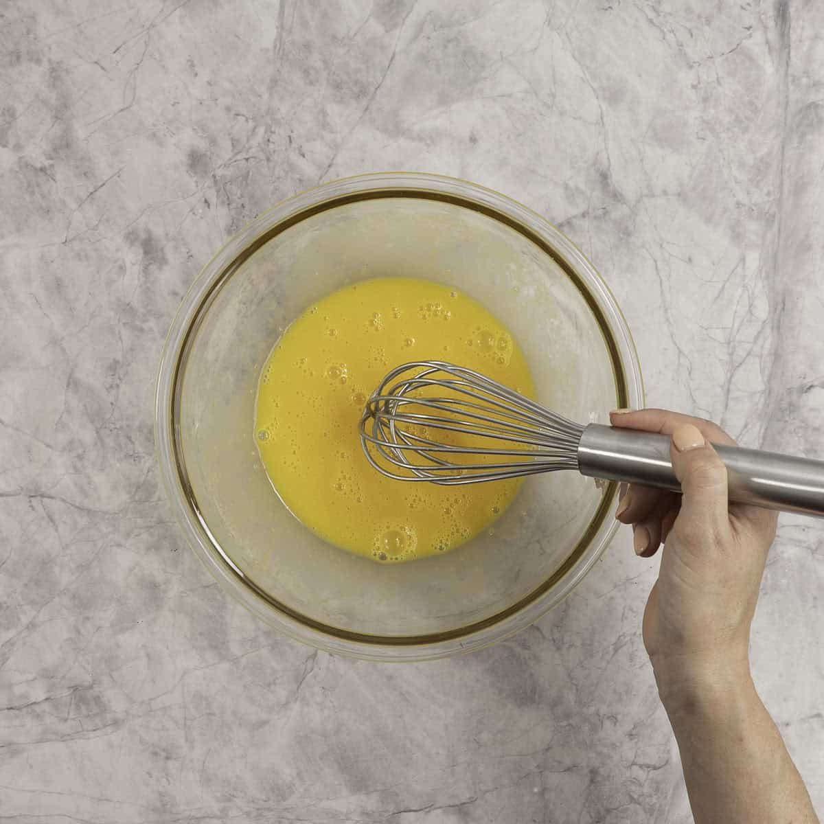 Womans hand using a whisk to stir eggs together in glass bowl.