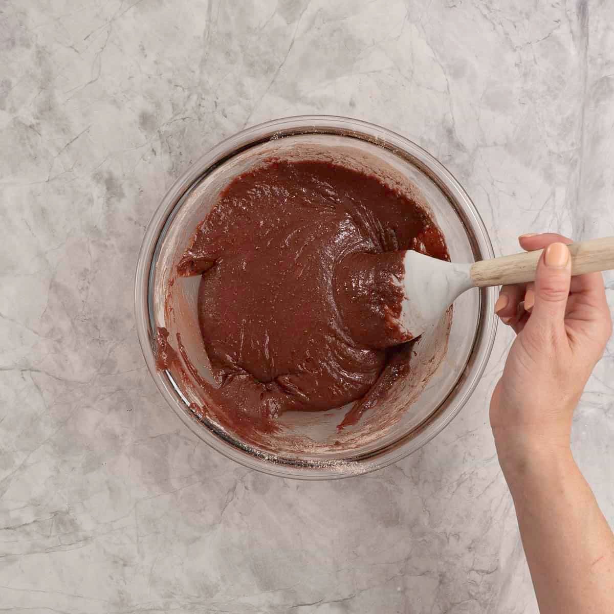 Batter for cookies in a large glass bowl with woman's hand mixing with a spatula.