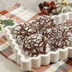 Red coloured cookies with cracked outta shell, coated in icing sugar placed in a white dish on christmas decorations in background and red and green checkered tea towel below.