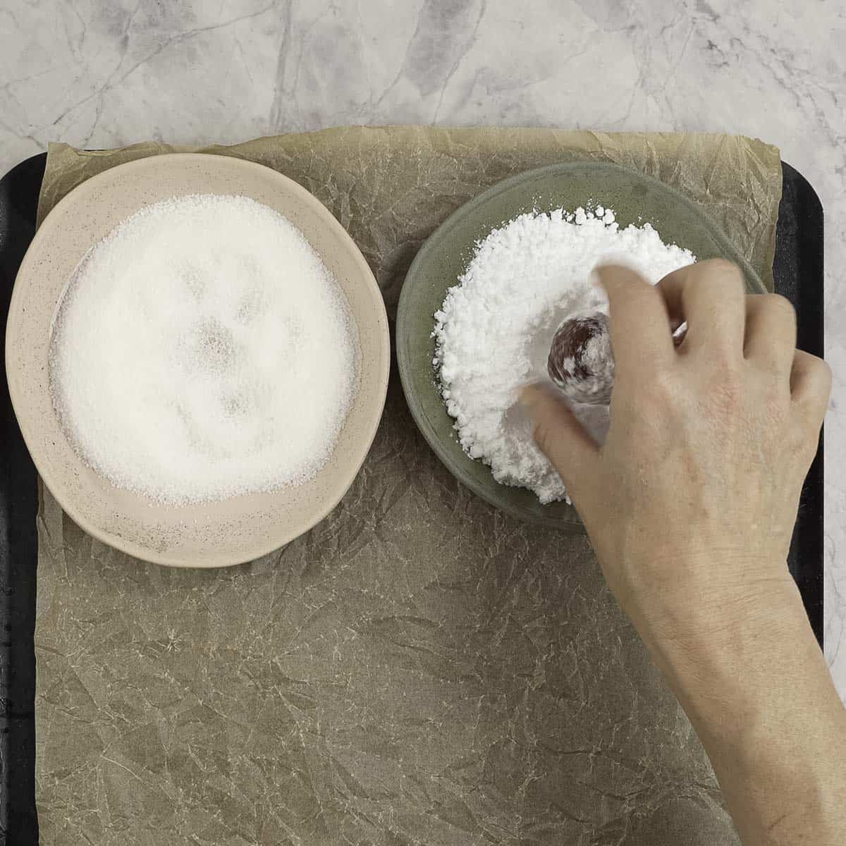 Two bowls one with sugar the other icing sugar, woman's hand coating cookie ball in icing sugar.