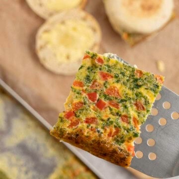 Metal spatula holding a square slice of sheet pan egg above a chopping board with breakfast muffins.