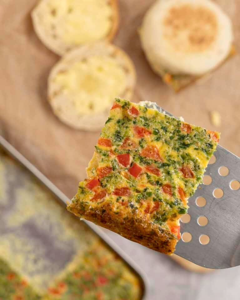 Metal spatula holding a square slice of sheet pan egg above a chopping board with breakfast muffins.