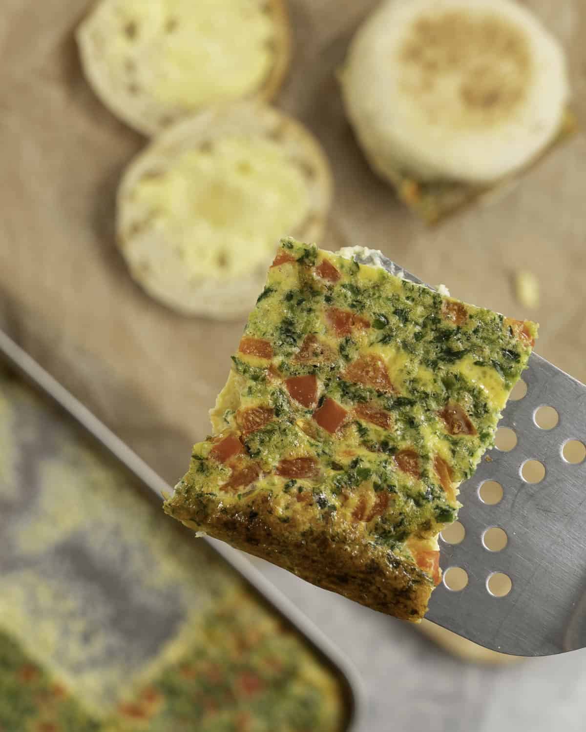 Metal spatula holding a square slice of sheet pan egg above a chopping board with breakfast muffins.