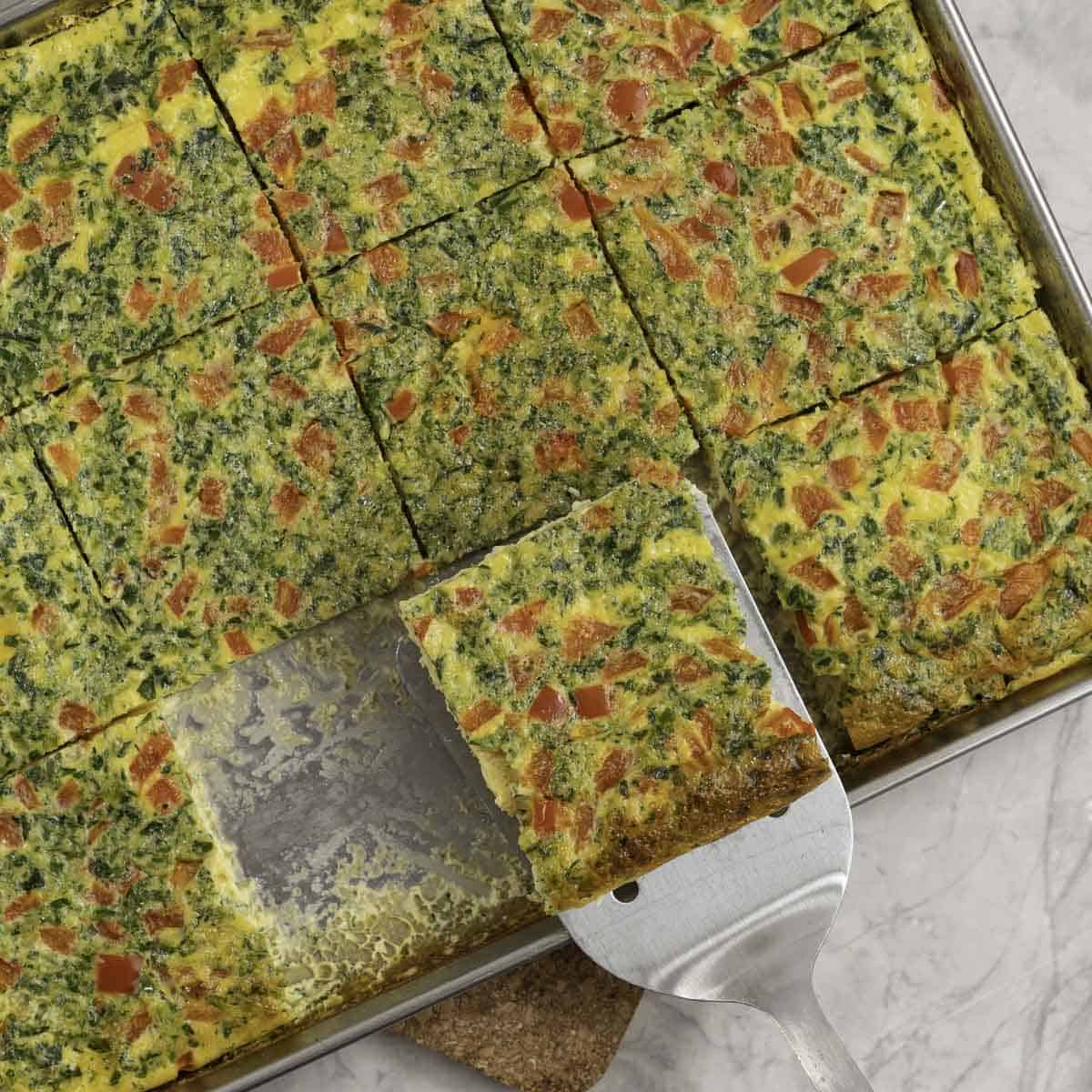 Sheet pan with eggs and vegetables cooked and cut into square pieces, metal spatula taking a slice out of tray.