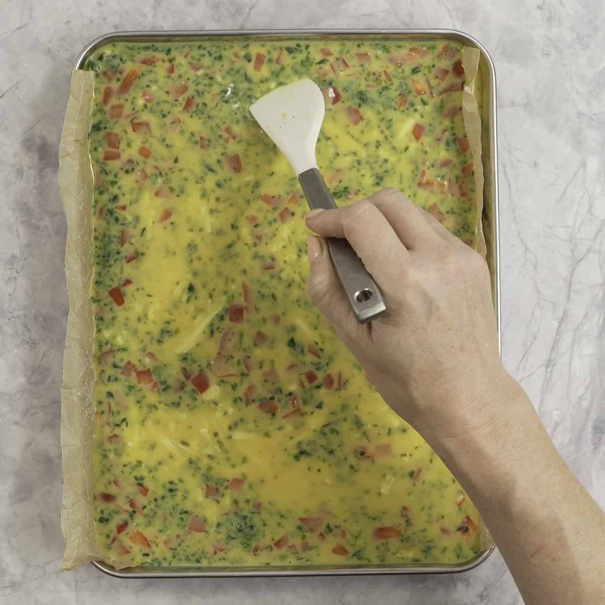 Sheet pan with eggy vegetable mixture inside, woman's hand holding a spatula moving ingredients around.