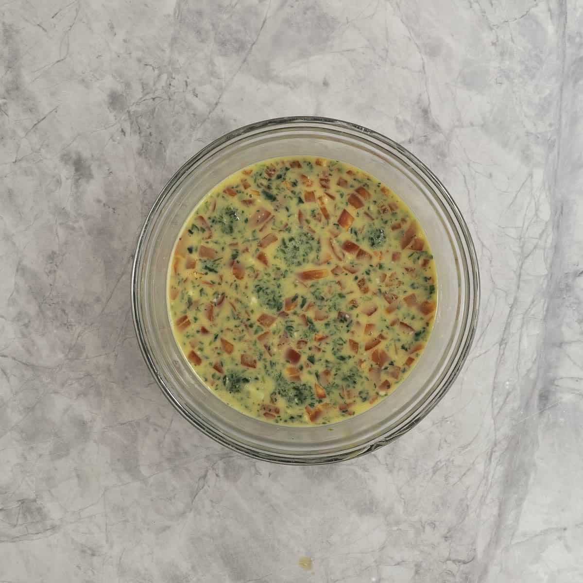 Diced capsicum, cheese and spinach mixed through eggy mixture in glass bowl on marble benchtop.