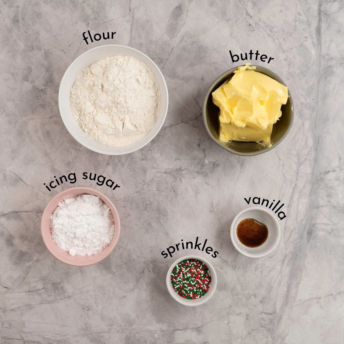 Bowl of flour, butter, vanilla, red and green sprinkles and icing sugar laid out on a bench top with text overlay